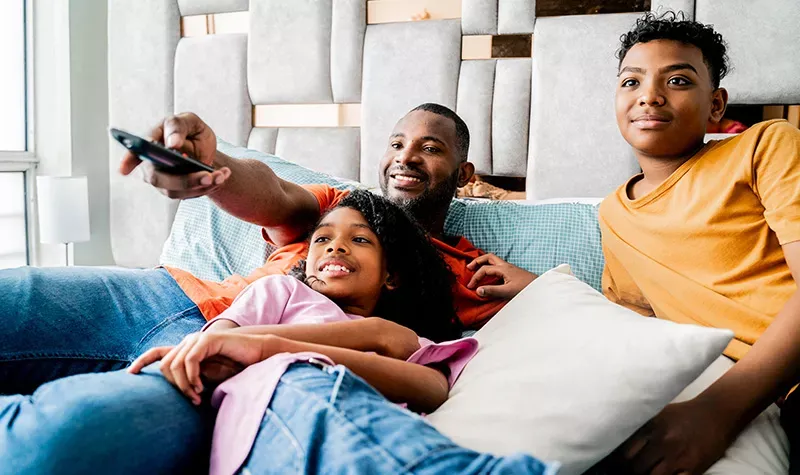 Father using remote control sits with children on couch watching TV